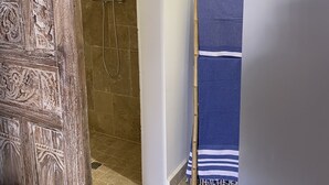 A bathroom with a shower, a wooden door with carvings, a towel rack with a blue towel, and a bamboo mat. - L'Oasis de Saint-Jacques (SAINT DENIS)
