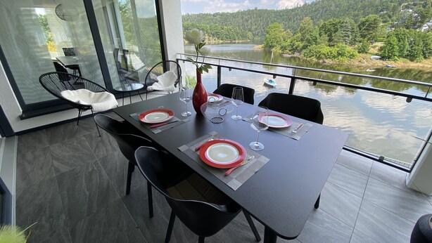 A dining area with a table set for two, overlooking a lake with a forested hillside in the background. - Apartments -Saaldorf WG 5 with balcony and view of the reservoir (Bad Lobenstein)
