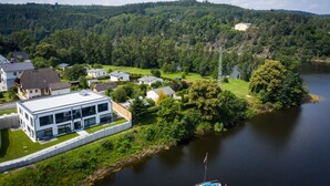 A modern building with a flat roof and large windows, surrounded by lush greenery and a lake. - Apartments -Saaldorf WG 5 with balcony and view of the reservoir (Bad Lobenstein)