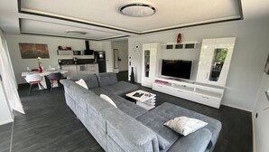 A modern living room with a grey sectional sofa, a flat-screen TV, and a dining area with a white table and chairs. - Apartments -Saaldorf WG 5 with balcony and view of the reservoir (Bad Lobenstein)