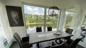 A modern dining area with a large table, chairs, and a view of a lake and greenery. - Apartments -Saaldorf WG 5 with balcony and view of the reservoir (Bad Lobenstein)
