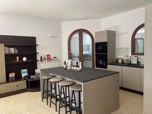 Una cuina moderna amb una illa central, tamboretes de bar, una prestatgeria i una finestra amb persianes.