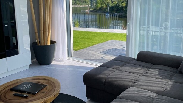Un salon moderne avec un grand canapé gris, une table basse en bois et un téléviseur posé sur un meuble blanc. Une grande fenêtre offre une vue sur un lac et une forêt.
