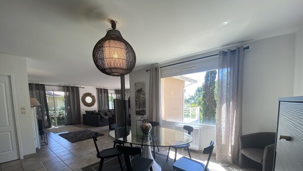 Una sala de estar moderna con una mesa de comedor de vidrio, sillas negras, una lámpara colgante trenzada grande y vistas de un jardín a través de una puerta corrediza de vidrio.