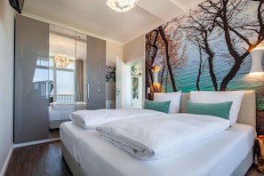 A bedroom with a large bed, a wardrobe, a painting of trees, and a view of the sea.