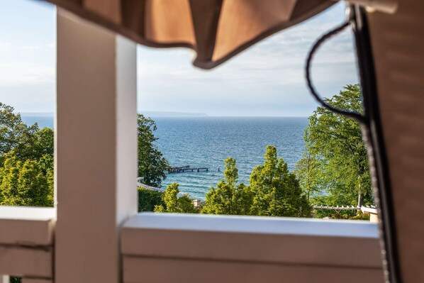 A view through a window with a curtain, overlooking a body of water, trees, and a pier. - Fantastic sea view from the balcony, use of swimming pool and sauna - Villa Bergfrieden (Göhren)
