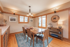 A dining area with a wooden table, chairs, and a chandelier. There are windows with blinds, a clock on the wall, and a lamp on a console table.