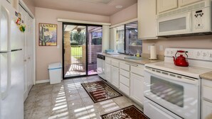 A kitchen with white cabinets, a microwave, a stove, and a red kettle. There is a door leading to a balcony with a garden view. - Corsican Villas 2BD Condo- Near Living Desert (Palm Desert)