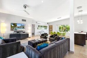 A modern living room with a large flat-screen TV, dark sofas, a coffee table, and a dining area with green chairs.