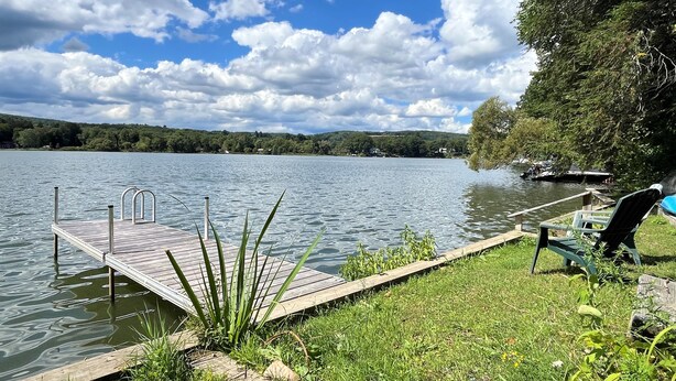 Un muelle de madera que se adentra en un lago tranquilo, con una zona de césped y una silla a la derecha.