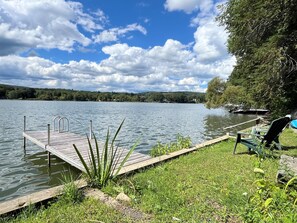 Eine Holzstege ragt in einen ruhigen See hinein, mit einer Rasenfläche und einem Stuhl auf der rechten Seite.