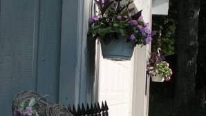 Una casa de paredes blancas, cerca de metal negro y macetas colgantes con flores moradas.