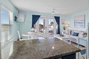 A spacious living area with a large granite island, a sofa with striped cushions, a dining table with chairs, and a view through a glass door to an outdoor patio.