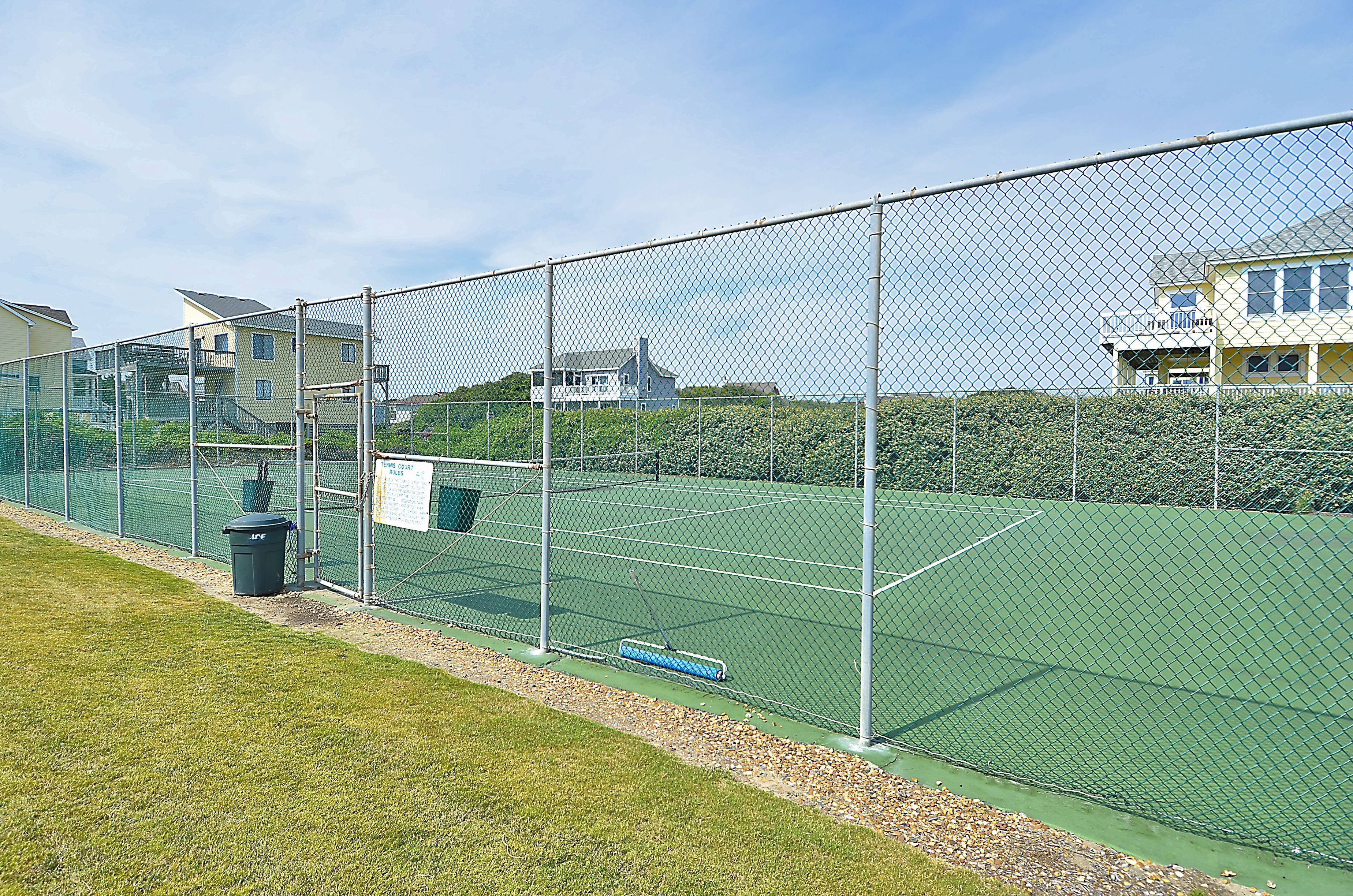Ein eingezäunter Tennisplatz mit grüner Belag, ein Mülleimer und ein blaues Objekt am Boden.