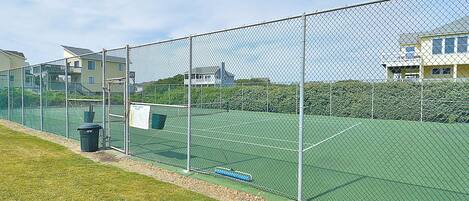 Ein eingezäunter Tennisplatz mit grüner Belag, ein Mülleimer und ein blaues Objekt am Boden.