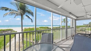 A balcony with a glass balustrade overlooking a beach, furnished with a table and chairs, and a ceiling fan. - Sanibel Surfside 115: Gorgeous Gulf Front Oasis! (Sanibel)