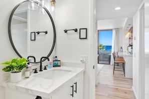 A modern bathroom with a large mirror, a white sink, and a potted plant. - Summer Deal Ocean View Corner Condo at Magic Sands Beach Kona (Kailua-Kona)