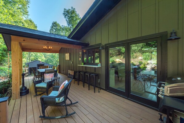 Una terraza de madera con mobiliario exterior, una zona de bar y amplios ventanales correderos que ofrecen vistas a un paisaje natural.
