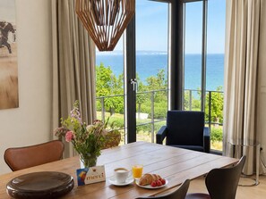 Uma área de refeições com mesa, cadeiras, um vaso de flores, uma xícara de café e um prato com croissant, com vista para a paisagem marítima.