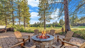 Ein Steine Feuerkorb mit vier hölzernen Adirondack-Stühlen darum, auf einer Ziegelterrasse gelegen, mit Blick auf einen Wald und einen Golfplatz im Hintergrund.