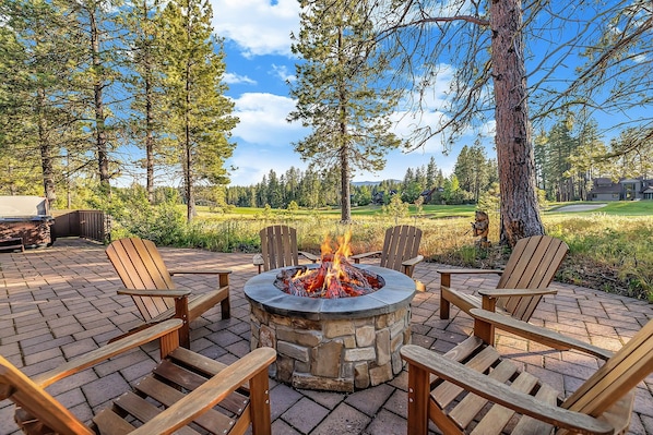 A stone fire pit with four wooden Adirondack chairs around it, set on a brick patio with a view of a forest and a golf course in the background.