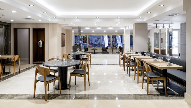 A modern dining area with round tables and chairs, a long bench, and a view of the street through large windows. - Merlin Hotel Istanbul (Istanbul)