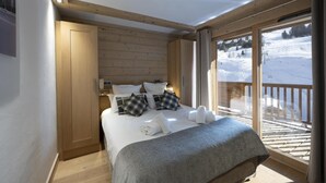 Habitación de hotel con decoración de madera, una cama grande con ropa blanca, vista a un paisaje nevado a través de un ventanal amplio y un balcón con barandilla de madera.