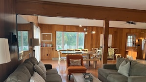 A cozy living room with a wooden interior, a dining area with a table and chairs, and a large window offering a view of the outdoors. - E-SEA-LIVIN' Oceanside Coastal Cottage in Corolla, 4 min walk to the beach! (Corolla)