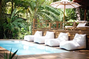 Área de piscina com deck de madeira, cadeiras de lounge acolchoadas em branco e um guarda-sol.
