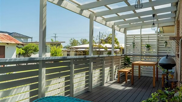 A covered outdoor deck with a wooden table and chairs, a hanging light fixture, and a view of surrounding buildings.