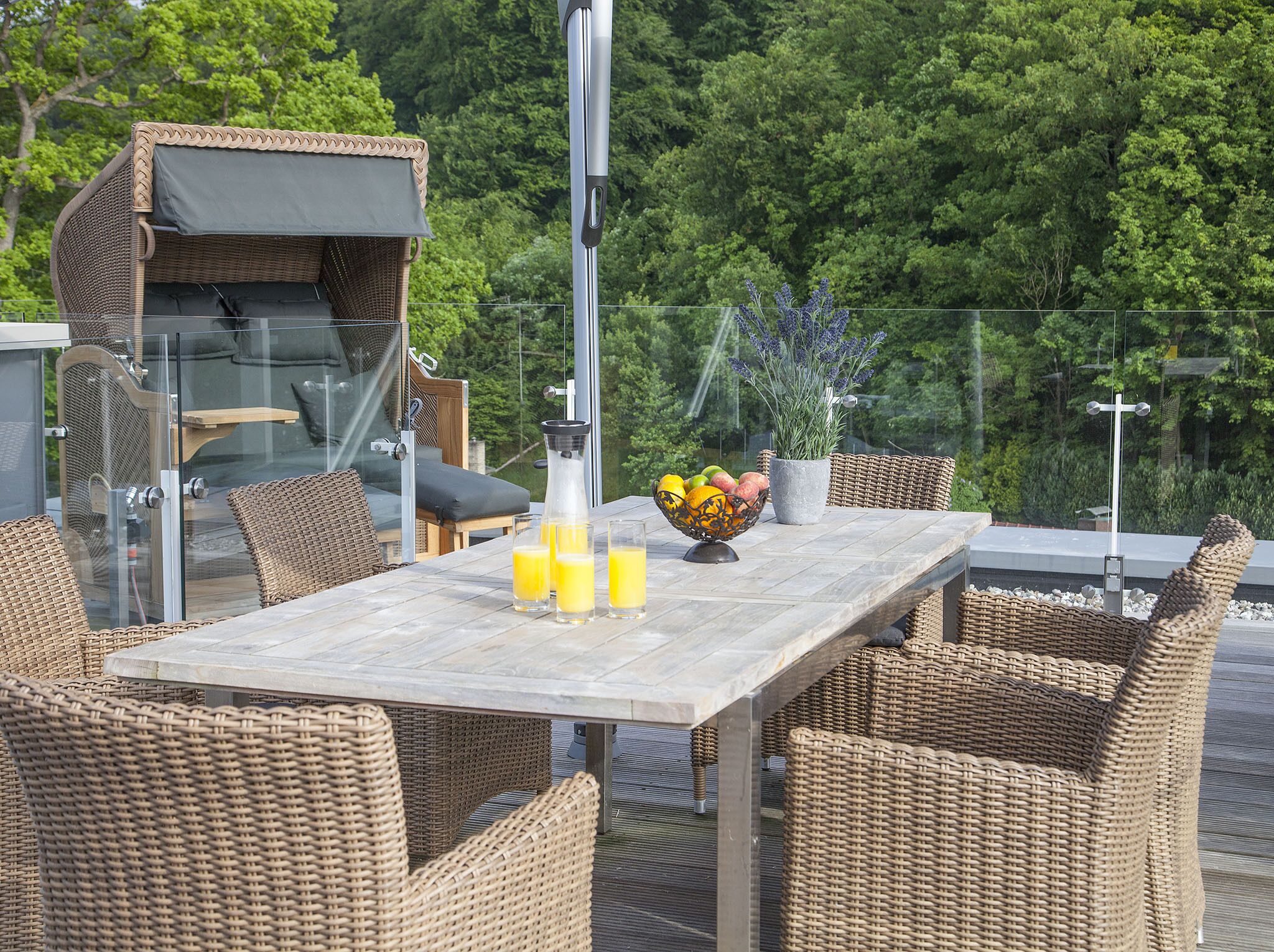 An outdoor dining area with wicker furniture, a table with a bowl of fruit and glasses of orange juice, and a grill in the background.
