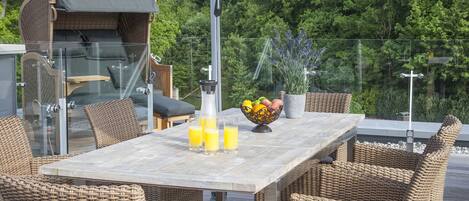 An outdoor dining area with wicker furniture, a table with a bowl of fruit and glasses of orange juice, and a grill in the background.