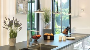 Une cuisine moderne dotée d’une grande fenêtre, de luminaires suspendus, d’un évier et d’un plan de travail sur lequel se trouve un saladier rempli de fruits et une corbeille de pain.
