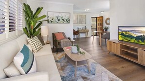 Un salon moderne avec un canapé, une table basse et un téléviseur. Il y a un coin repas avec des chaises et une table. Une grande plante se trouve dans un coin.