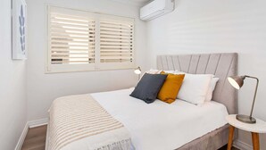 Un lit bien fait, recouvert d’une couverture à rayures, une lampe de chevet et une fenêtre avec des stores.