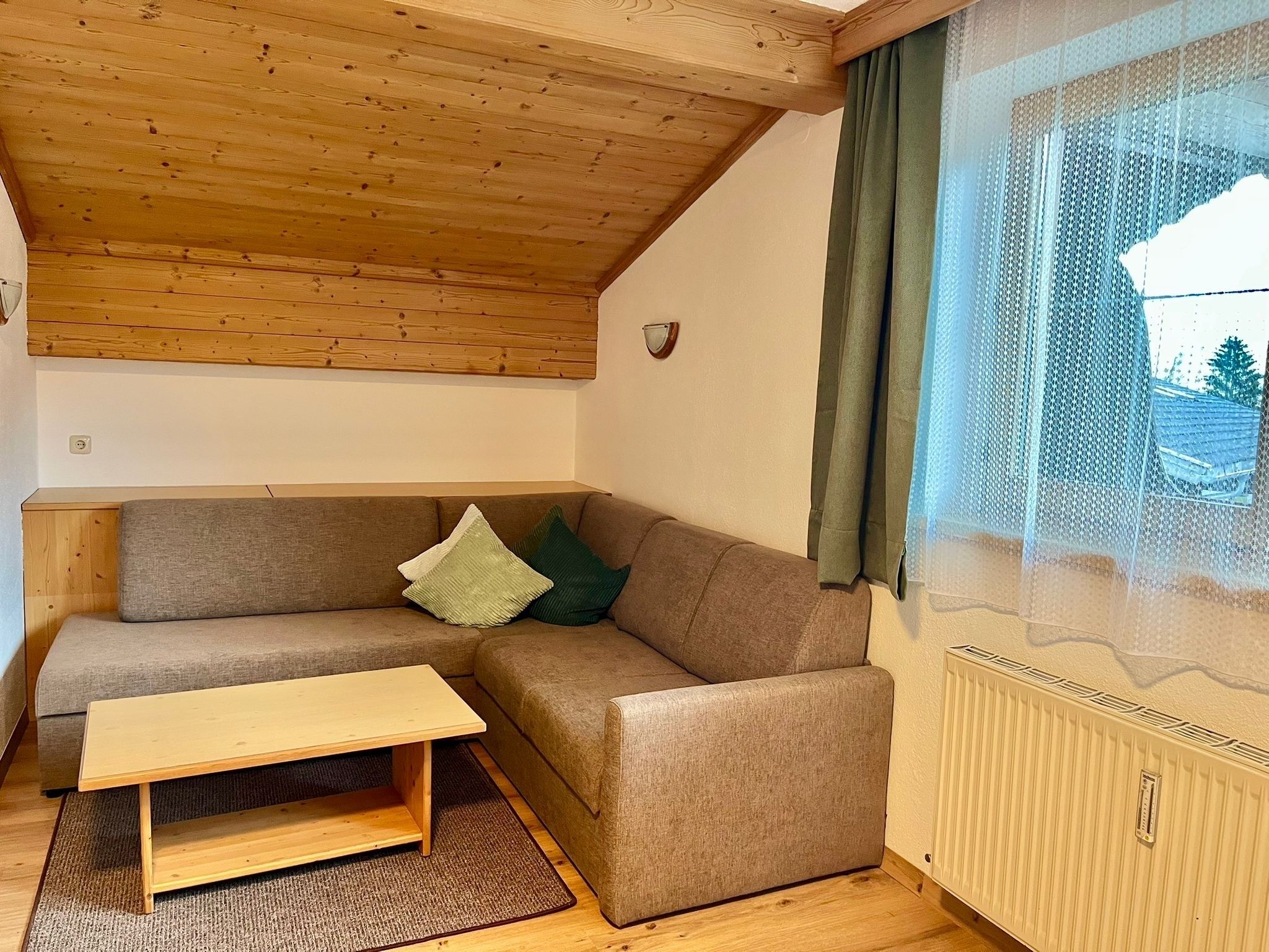 Una habitación acogedora con techo de madera, un sofá marrón con cojines, una mesita de madera y una ventana con vistas a un edificio.