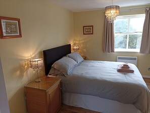 Een netjes opgemaakt bed met hoofdbord, nachtlampjes en een raam met gordijnen.