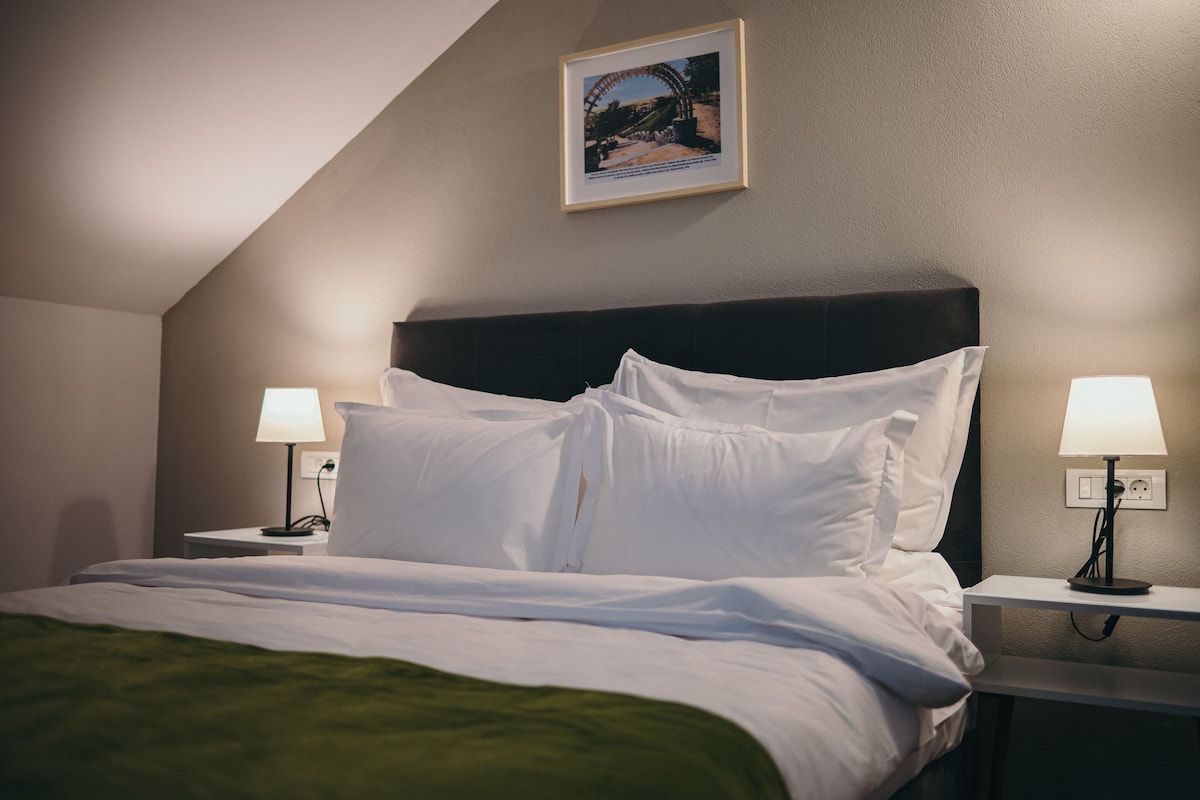 Ein ordentlich bezogenes Bett mit weißen Leinen, zwei Nachttische mit Lampen und ein gerahmtes Bild an der Wand.