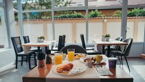 A breakfast table set with eggs, sausages, a hot dog, and a drink, with a view of a garden through large windows. - NEST Guesthouse (Drobeta-Turnu Severin)
