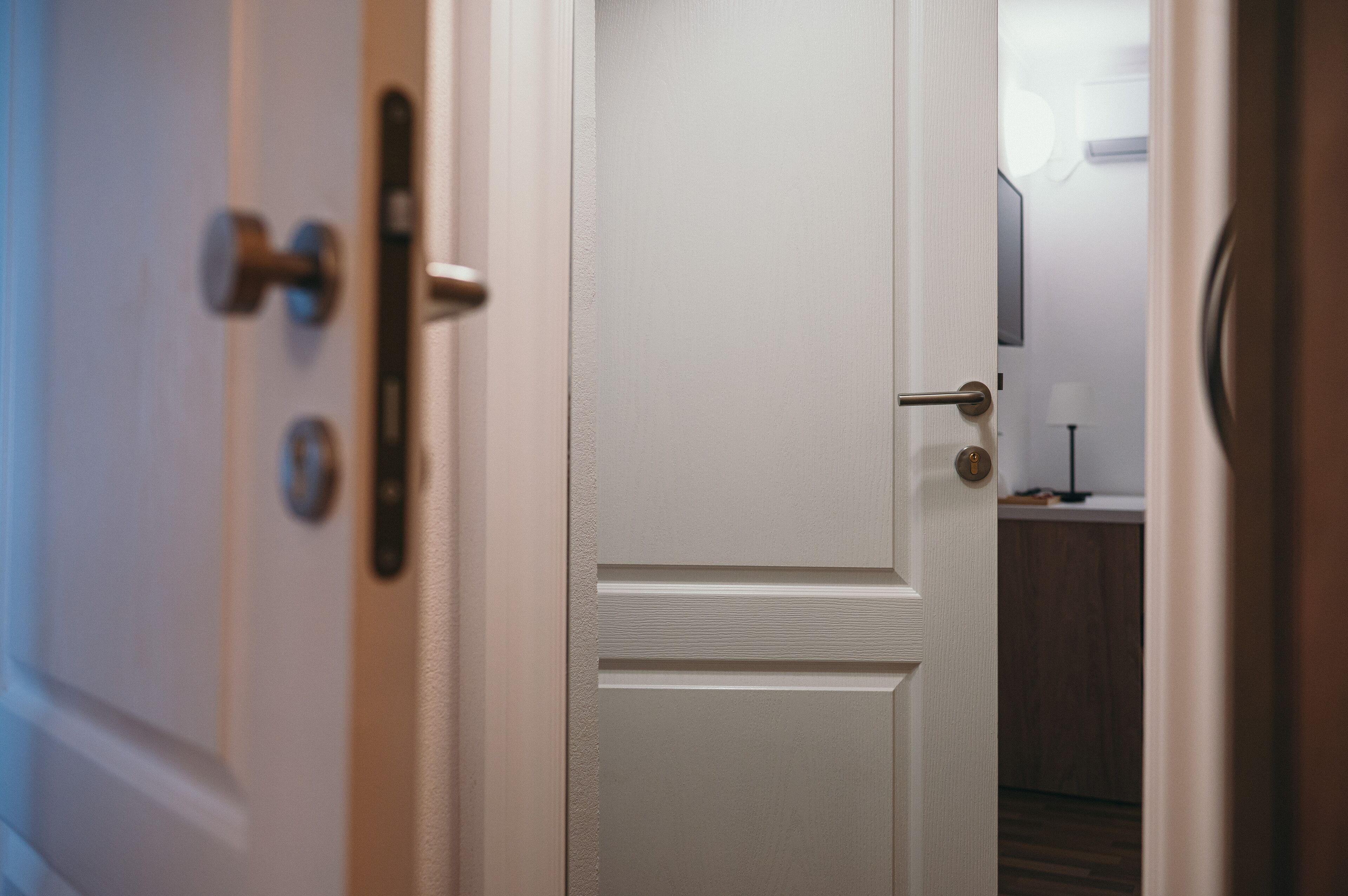 A white door with a handle and a lock, leading to a room with a wooden floor and a small table with a lamp.