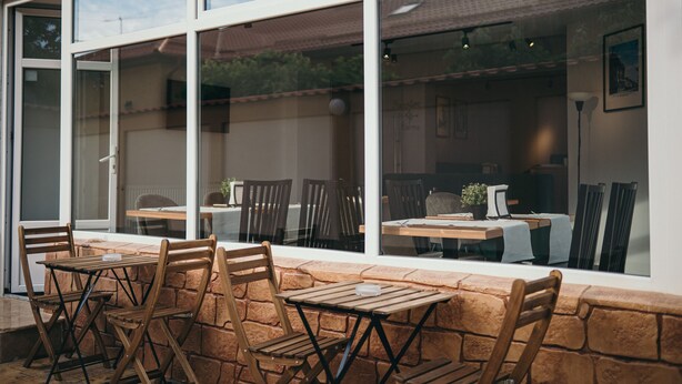 An outdoor patio with wooden tables and chairs, large windows, and a stone wall. - NEST Guesthouse (Drobeta-Turnu Severin)