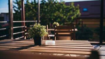 Una mesa de madera con una planta en maceta y un recipiente de plástico transparente en un balcón con baranda, con vistas a un edificio y árboles.