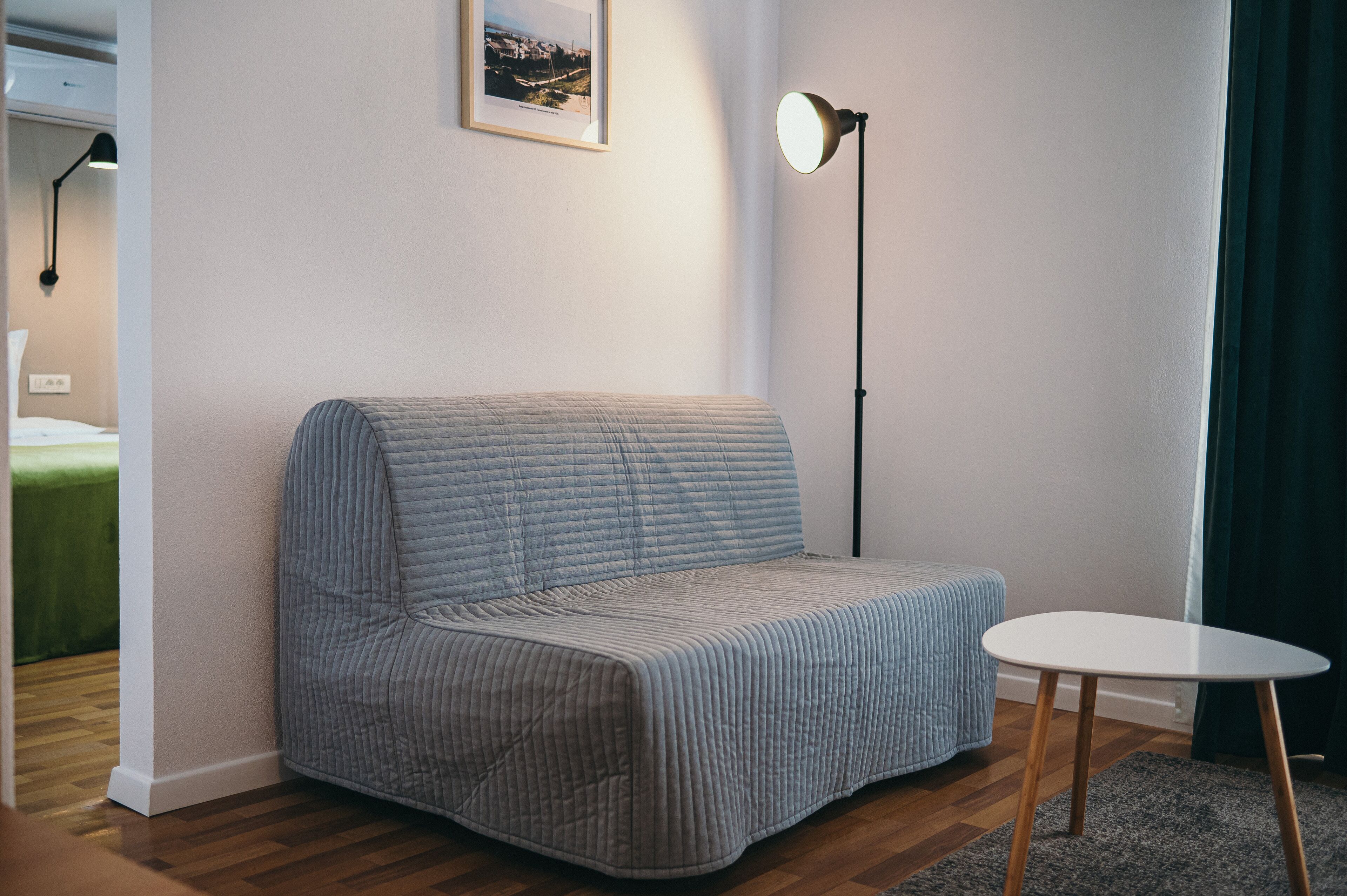 Una habitación con un sofá gris, una mesita blanca, una lámpara de pie y un cuadro en la pared.