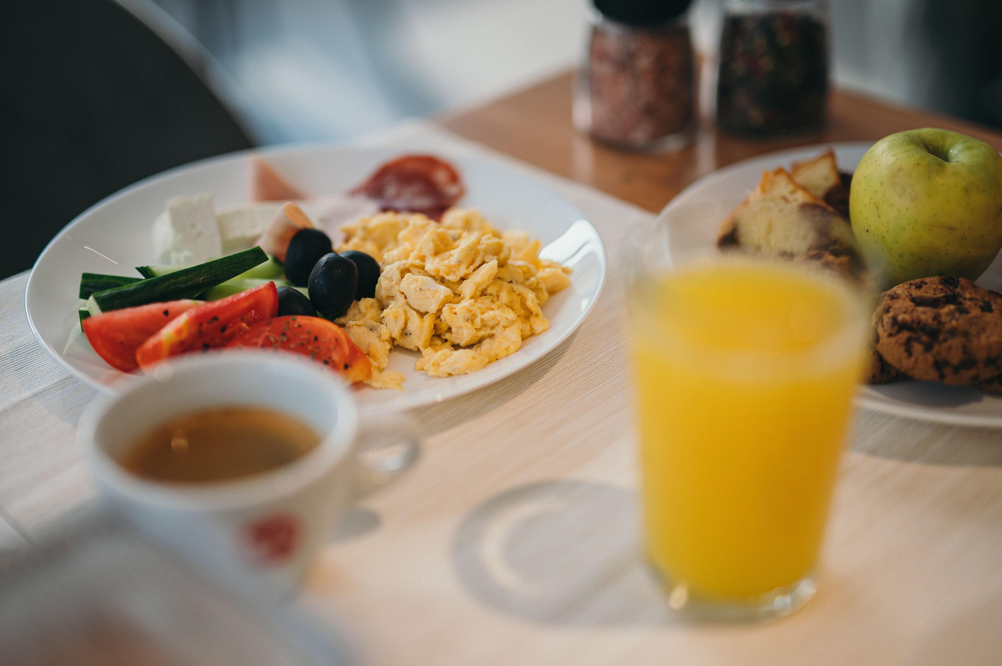 A breakfast setting with plates of scrambled eggs, tomatoes, black olives, and a green apple. There is a glass of orange juice and a cookie on the table.