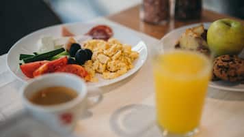 A breakfast setting with plates of scrambled eggs, tomatoes, black olives, and a green apple. There is a glass of orange juice and a cookie on the table.