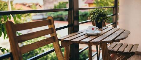 A wooden table with a white cup and saucer, a potted plant, and a wooden chair on a balcony with a view of greenery and buildings.