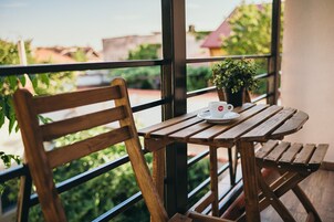Uma mesa de madeira com uma xícara e pires brancos, um vaso com planta e uma cadeira de madeira em uma varanda com vista para áreas verdes e edifícios.