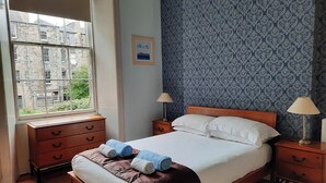 A neatly made bed with pillows and rolled towels, wooden nightstands with lamps, a window with a view of buildings, and a patterned wall. - Cornwall Street [297] is a Spacious and charming apartment close to Usher Hall (Edinburgh)