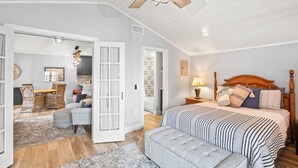 Een slaapkamer met een groot bed, een houten hoofdeinde, een plafondventilator en uitzicht op een andere kamer met een eethoek.