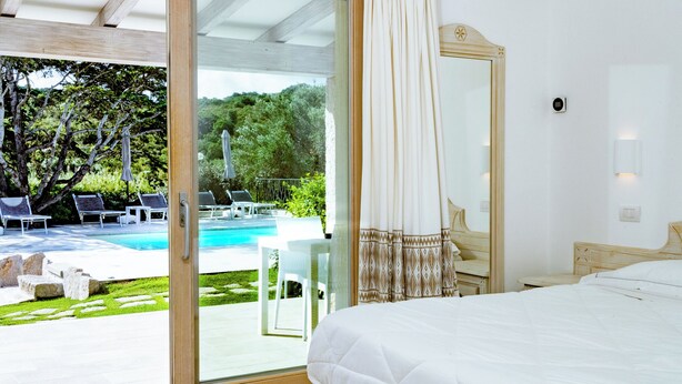 A bedroom with a large bed, a glass door leading to a pool area with lounge chairs and a view of greenery. - La Corte di Pulicinu (Arzachena)